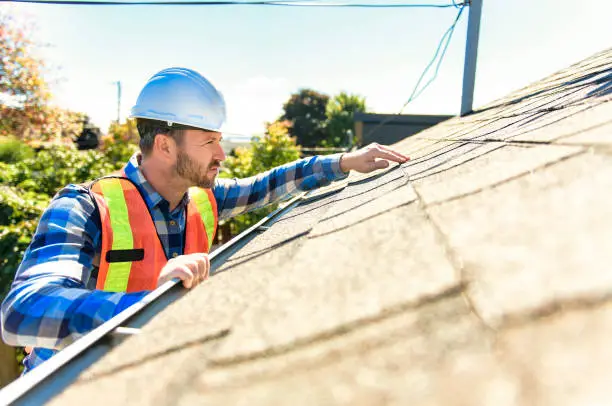 Roofing contractor inspecting asphalt shingles on a residential home roof during a sunny day.